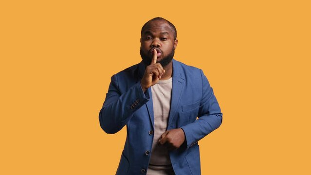 Annoyed african american man doing shushing hand gesturing, irritated by noise, having negative mood. Person placing finger on lips, doing quiet sign gesture, studio background, camera B