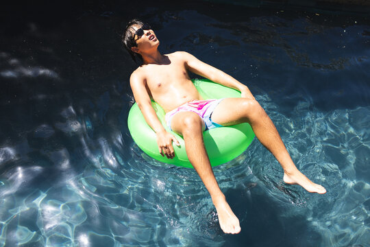 A teenage Asian boy wearing sunglasses relaxing on a green float in a pool outside at home