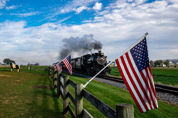 A vintage steam train chugs along the countryside, flanked by American flags on a wooden fence,...