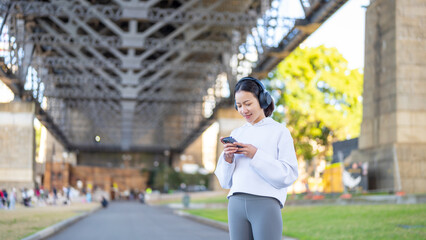 Asian woman in sportswear listening to music with headphones and mobile phone app jogging workout exercise at city street in the morning. Healthy care motivation sport training and running concept. - obrazy, fototapety, plakaty