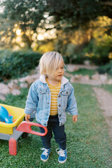 Little girl stands near a toy wheelbarrow in a park