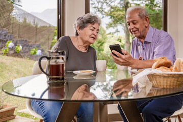 Elderly couples use a cell phone in the dining room of their house.