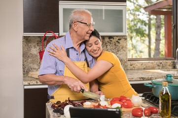 Daughter hugs her father tenderly in the kitchen while her father prepares food.