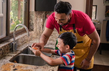 Latino father shows his son the correct way to use a sponge to wash dishes.