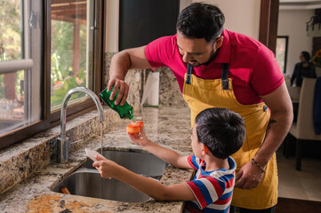 Latin father helps his son put soap on the sponge to wash the dishes. Father teaches his son.