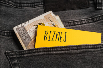 Yellow card with a handwritten inscription "Biznes", inserted into the pocket of gray pants jeasnow, next to Polish banknotes PLN (selective focus), translation: business