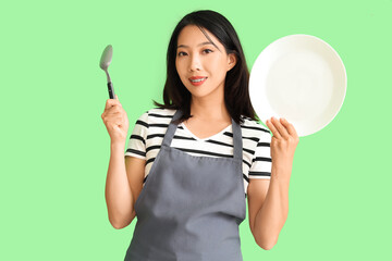Happy beautiful Asian woman with plate and spoon on green background