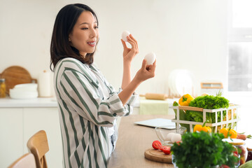 Beautiful young Asian woman with fresh eggs cooking in kitchen