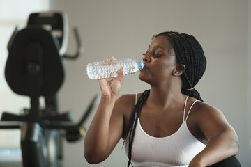 Athletic healthy strong female African American plus-size sportswear drinking water in bottle after...