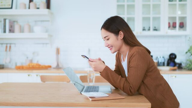 Happy young asian woman relax in domestic kitchen at home, using smartphone, smiling girl use cell phone chatting, browse wireless internet on gadget, shopping online from home