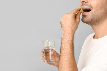 Young man with glass of water taking vitamin A pill on light background, closeup