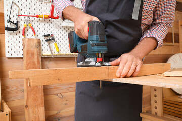 Mature carpenter sawing wooden board with jigsaw at table in workshop, closeup