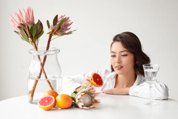 Young Asian woman with fresh grapefruit and beautiful protea flowers at table near white wall