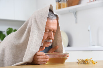 Mature man with towel doing steam inhalation at table in kitchen - obrazy, fototapety, plakaty