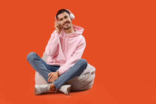 Handsome young man in headphones resting on beanbag chair and listening music against orange background