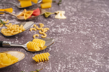Spoons with different uncooked pasta on grey grunge background, closeup