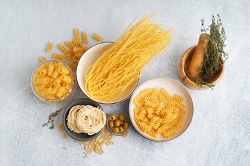 Bowls with different uncooked pasta, olives and thyme on light background