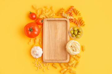 Different uncooked pasta, olives, tomatoes and wooden board on orange background