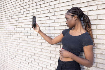 In this image, a young African woman is positioned in profile, holding up her smartphone to take a...