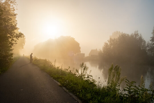 This serene image features a lone individual walking down a mist-covered riverside path, with the soft dawn light creating a halo through the trees. A house partially obscured by the mist can be seen