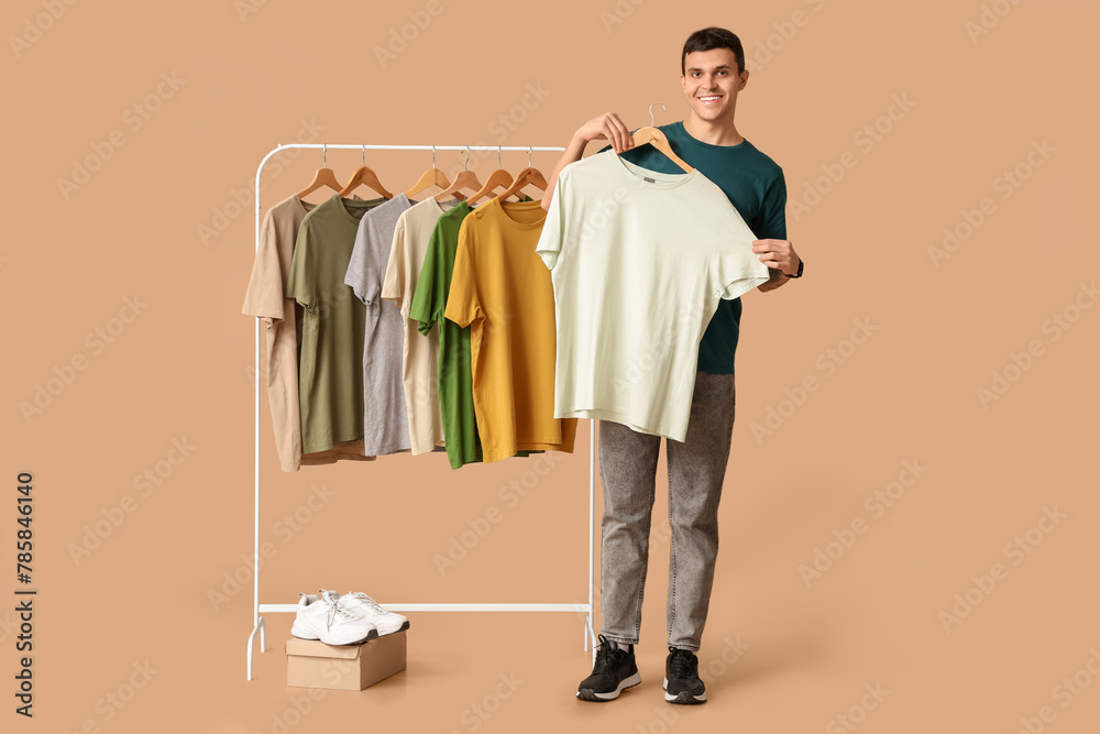 Wall mural Handsome young happy man and rack with different t-shirts on brown background