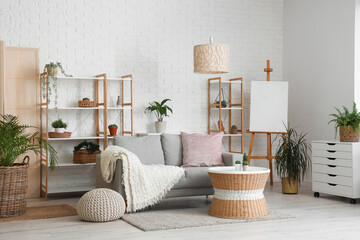 Loft style interior of light living room with cozy sofa, chest drawer, table, houseplants, shelving units and easel