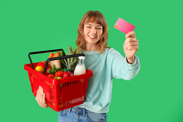 Young woman with full shopping basket and credit card on green background