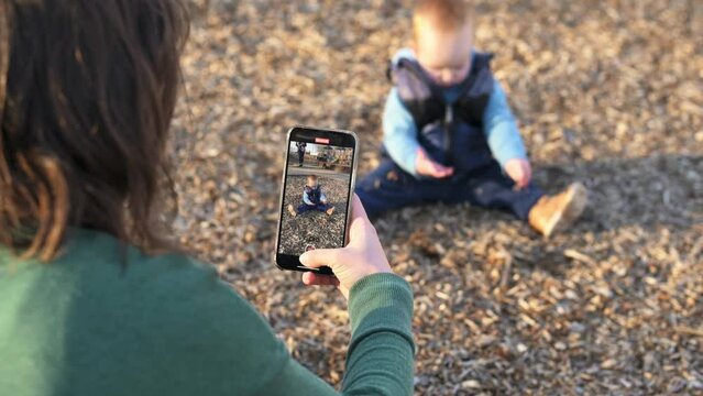 in the playground, the boy sits on the ground and plays with the ground, explores nature, the mother takes photos and videos on the phone for childhood memories