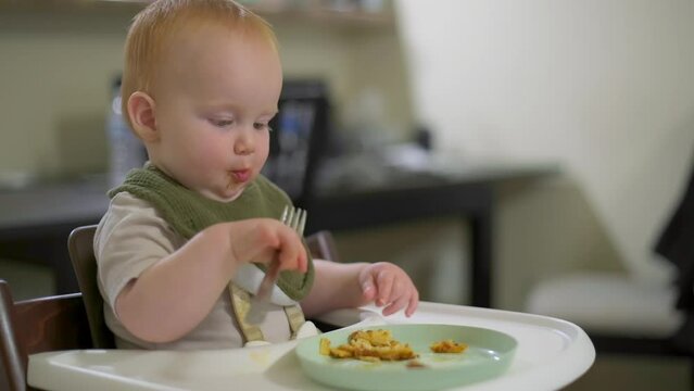 the one year old boy is sitting in the dining room and learning to eat with a fork himself