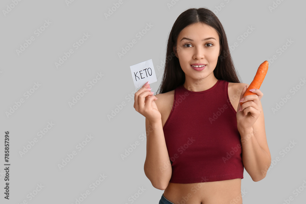 Wall mural young happy asian woman holding sticky note with word keto and carrot on grey background