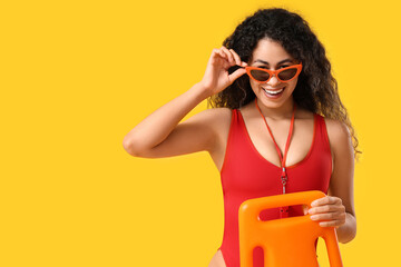 Beautiful young happy African-American female lifeguard with rescue buoy on yellow background
