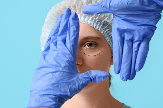 Plastic surgeon examining woman's face on blue background, closeup. Blepharoplasty concept - Powered by Adobe