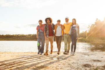 A bonded group of five strolls by the water's edge as the sun sets, casting a soft glow on their casual outing. Close-knit Friends Walking Together Along a Lakeshore at Dusk. High quality photo