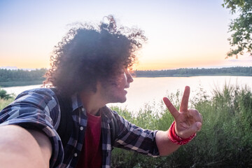 Person with curly hair making a peace sign, with a serene lake and sunset backdrop, radiating calm...