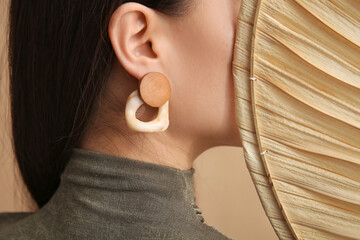 Young woman with beautiful earrings and bamboo fan on beige background