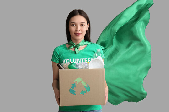 Young female volunteer in superhero cape holding recycle box with garbage on white background