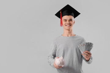 Male student in graduation hat with piggy bank and money on grey background. Concept of savings for...