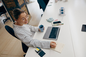 Young female entrepreneur working on laptop and making notes while sitting in stylish office 