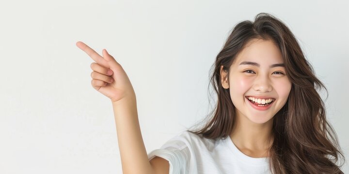 A young Asian woman wearing a white t-shirt stands isolated on a gray background, pointing her finger while smiling confidently and happily.