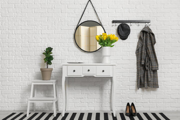 Vase of yellow tulips on dresser with mirror, hanger and houseplant near white brick wall in hallway