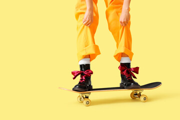 Stylish young woman in boots with bows on skateboard against yellow background, closeup