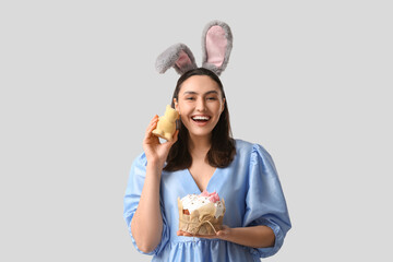 Happy young woman in bunny ears with sweet Easter cake and toy rabbit on grey background