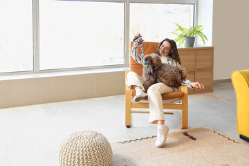 Young woman playing with cute poodle in armchair at home - obrazy, fototapety, plakaty