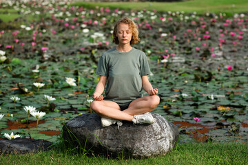 A young girl, with blond curly hair, meditates, sitting on a large stone, against the backdrop of...