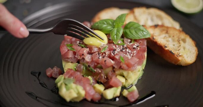 Tuna and avocado tartare with sesame seeds, capers and egg yolk on a dark ceramic plate with baguette bread crouton chips
