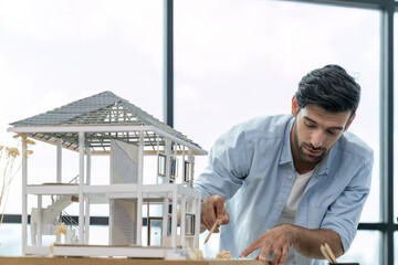 Caucasian architect engineer inspects house model while using pencil pointing at structure....