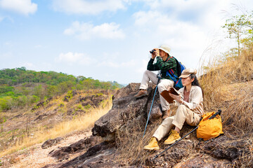 Two asian hiking backpacker sitting on the top of the mountain during a walk or surveying the forest in summertime,a man looking into binoculars and a woman looking away and taking notes on notebook