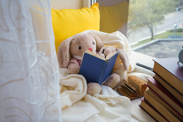 A toy hare is reading a book to a child sitting on a window in a white plaid