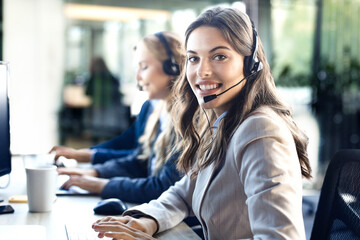 Female customer support operator with headset and smiling, with collegues at background. - 785936716