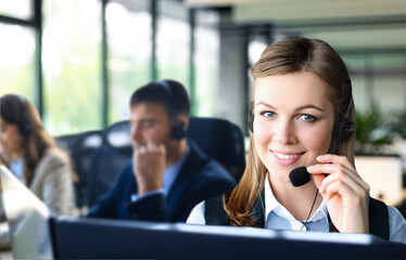 Female customer support operator with headset and smiling, with collegues at background. - 785936729